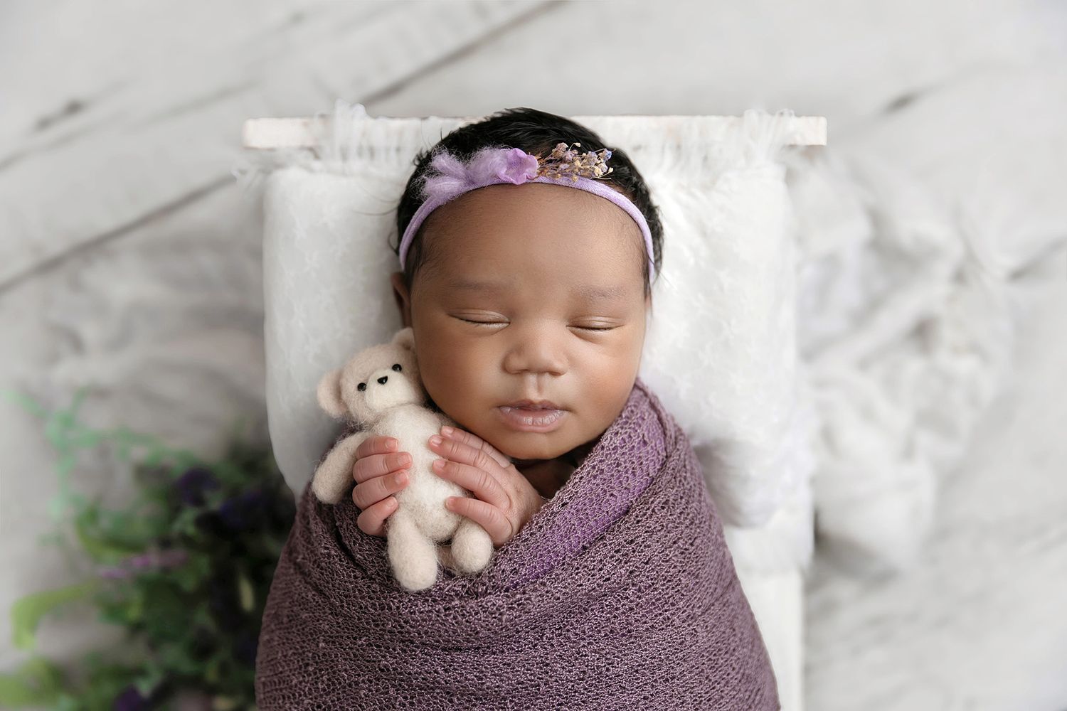 Image of baby girl in lilac purple lying in a white bed holding teddy.