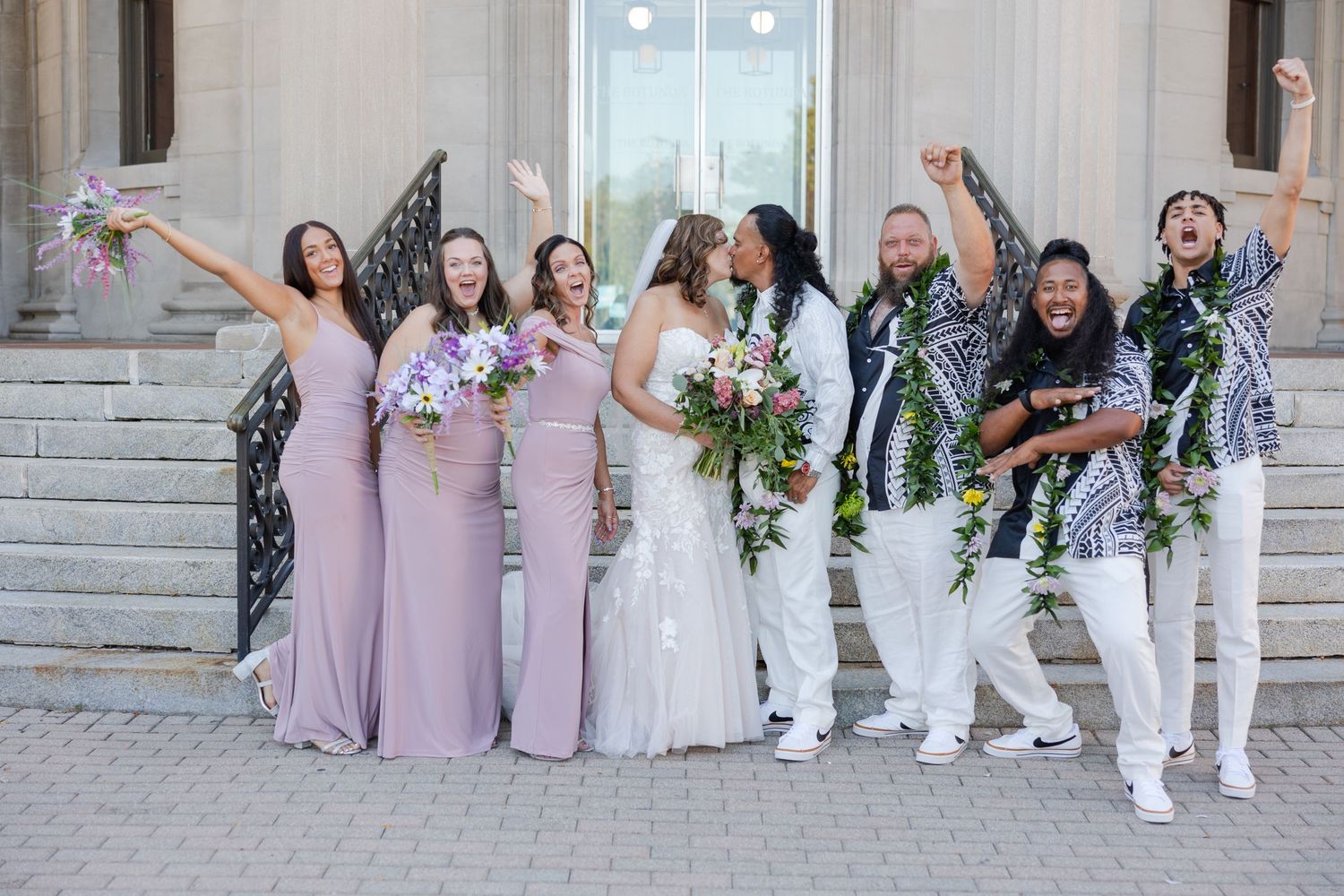 A bridal party surrounding the bride and groom cheering them on as they share a kiss.