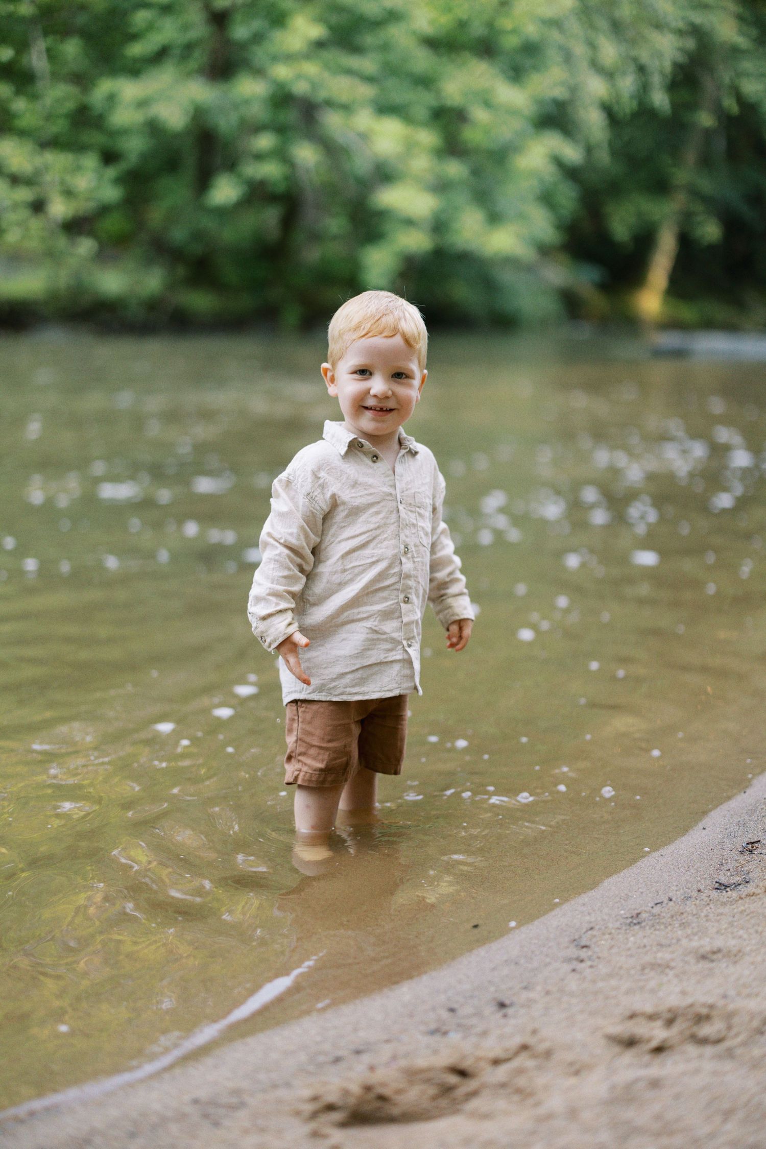 Young child wearing light colored shirt stands in shallow river water.