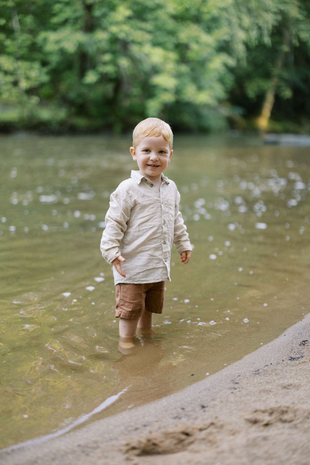 Young child wearing light colored shirt stands in shallow river water.