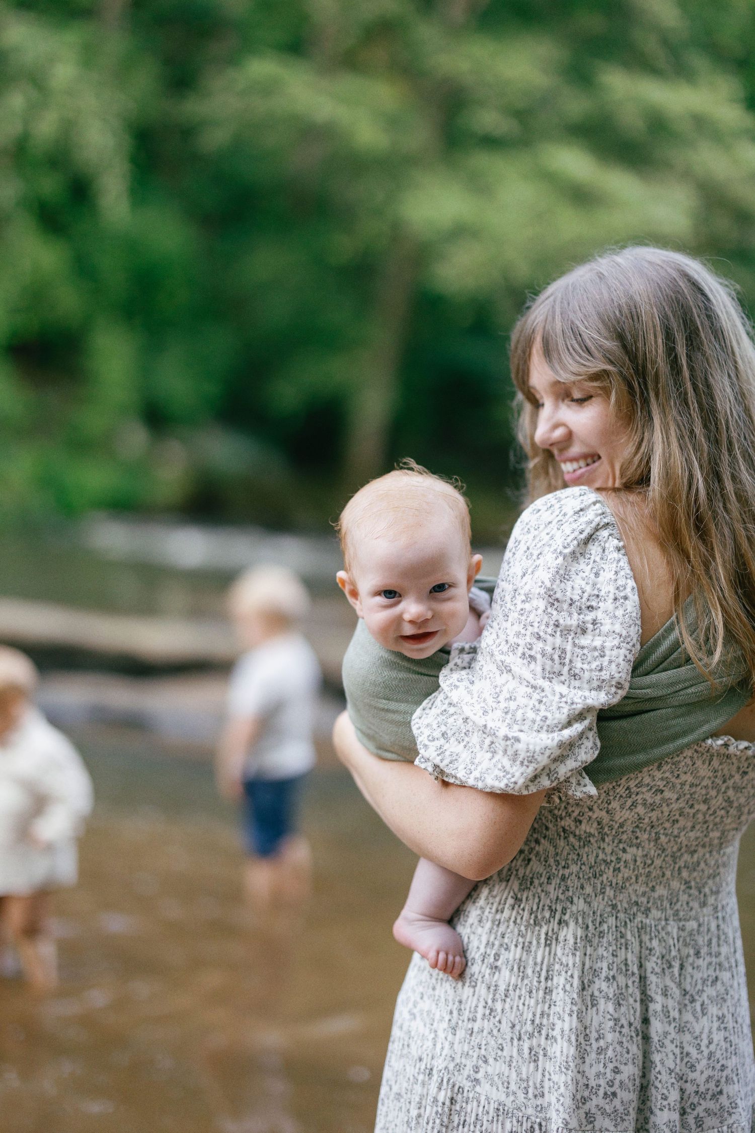 Parent wearing floral dress holds baby in carrier while standing by river.