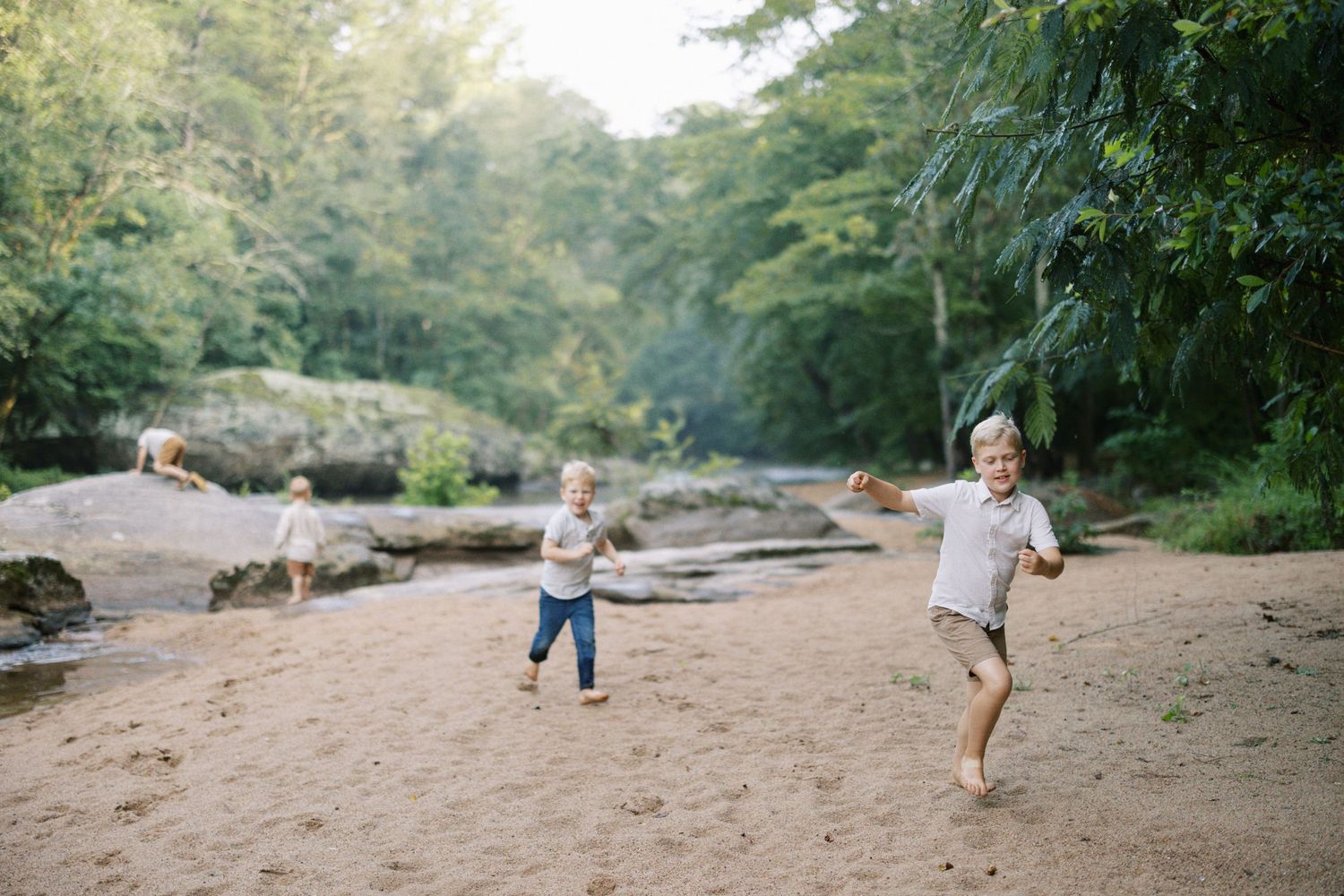 Two children running playfully on sandy riverbank with forest in background.