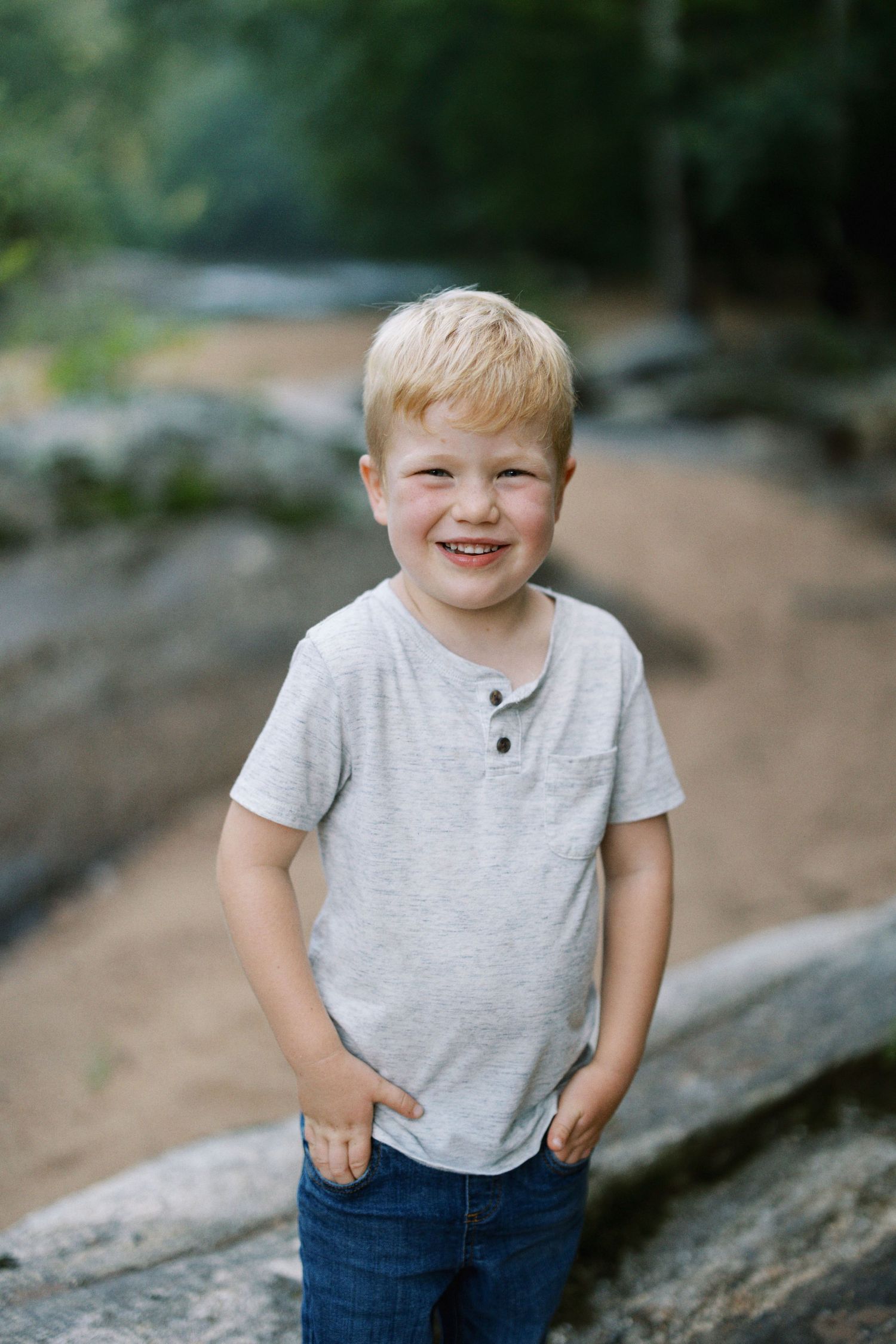 Young child in gray t-shirt poses outdoors on rocky trail with natural background.