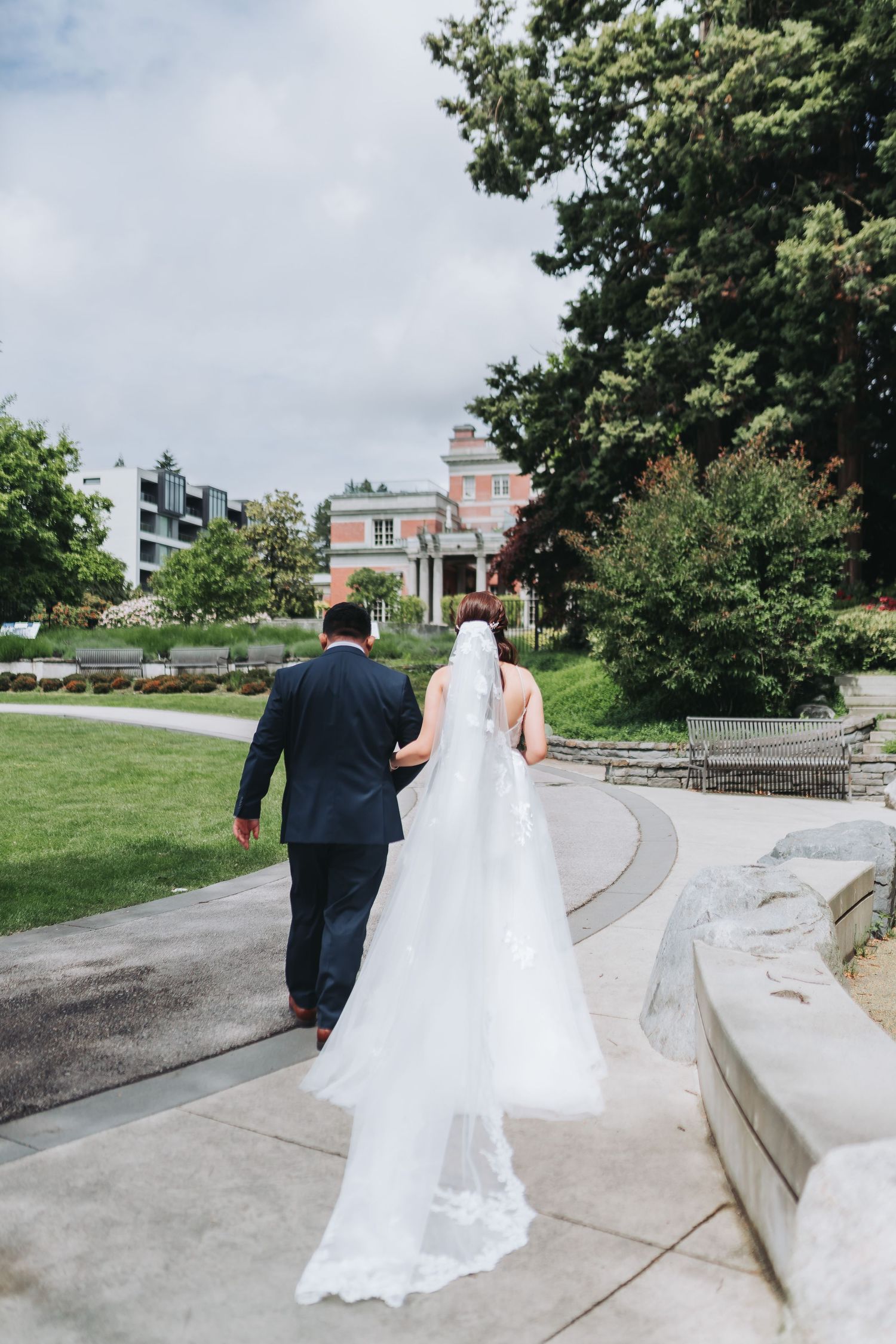 A couple in wedding attire walks along a curved garden path surrounded by lush greenery and buildings.