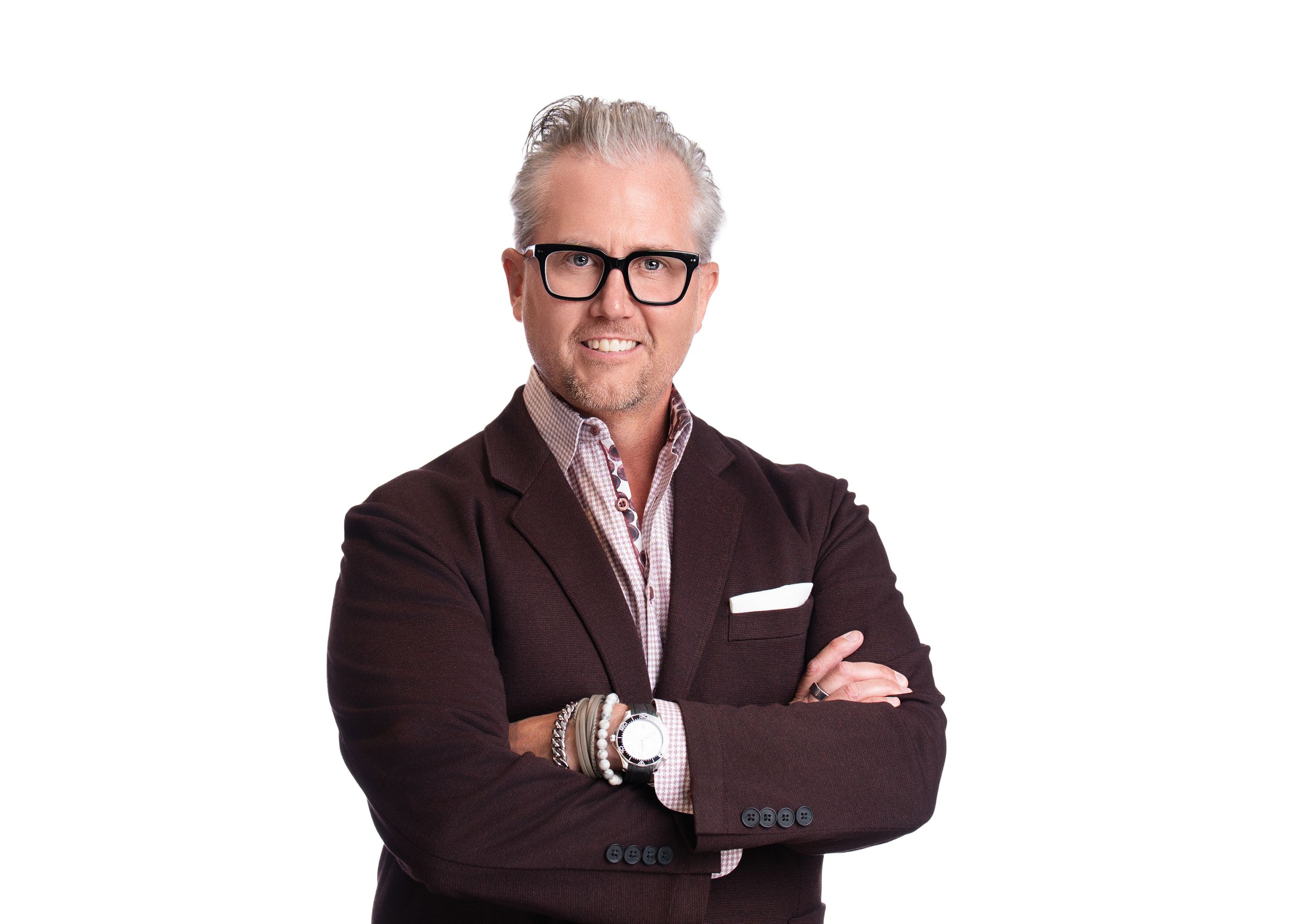 Professional portrait of someone wearing a brown blazer, white dress shirt, and black-rimmed glasses against white background.