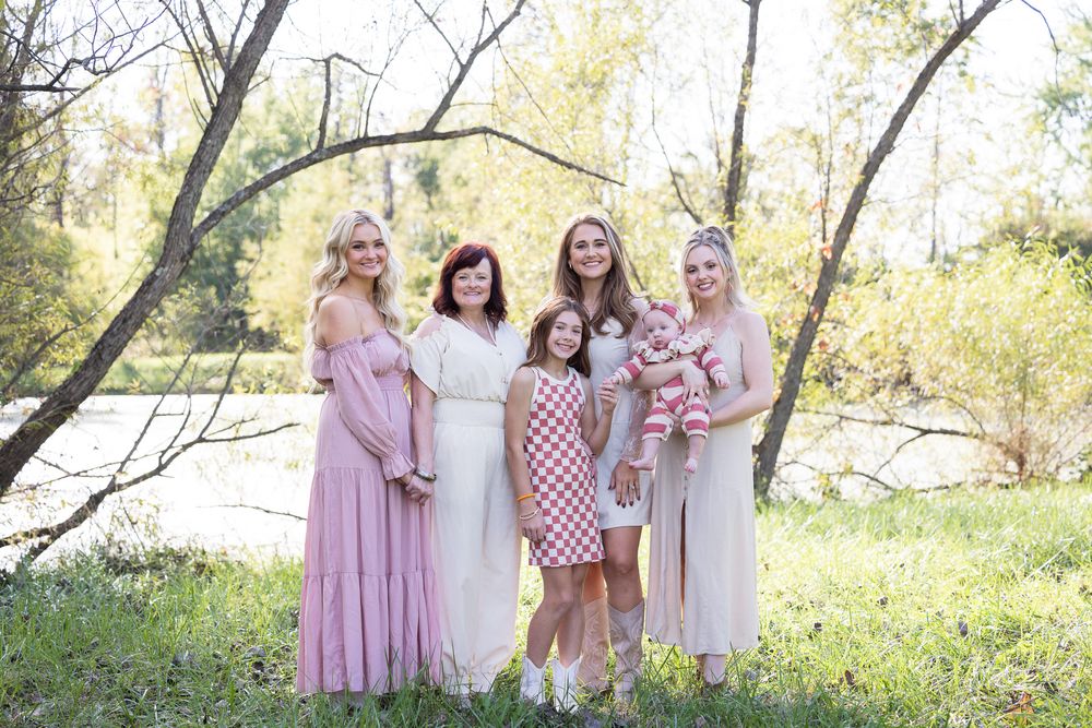 A group wearing coordinated pink and white dresses poses together outdoors by a lake.