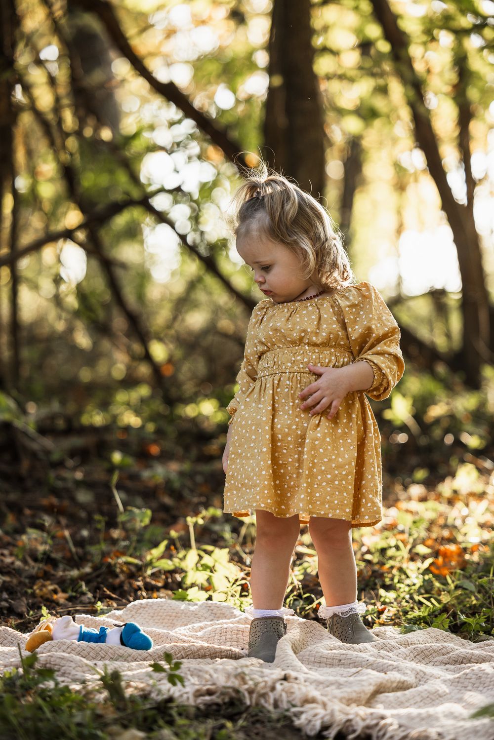 A toddler in a yellow polka dot dress stands on a blanket in a sunlit forest.