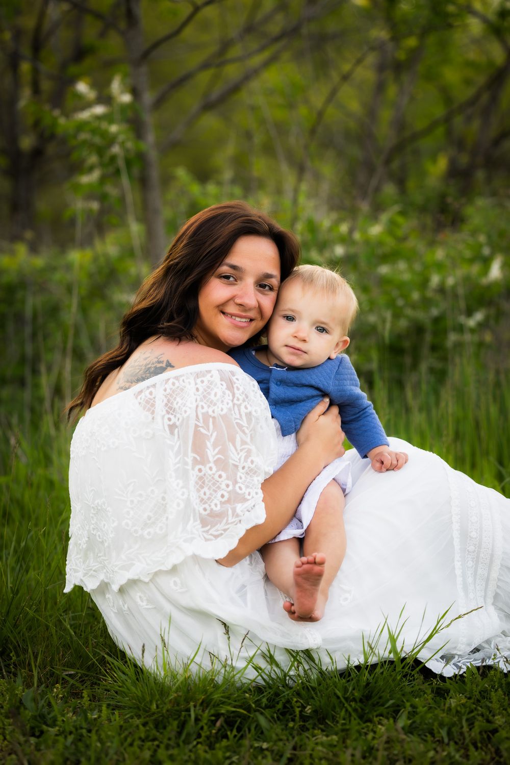 Mother in white lace dress holding baby in blue outfit while sitting in grassy field.