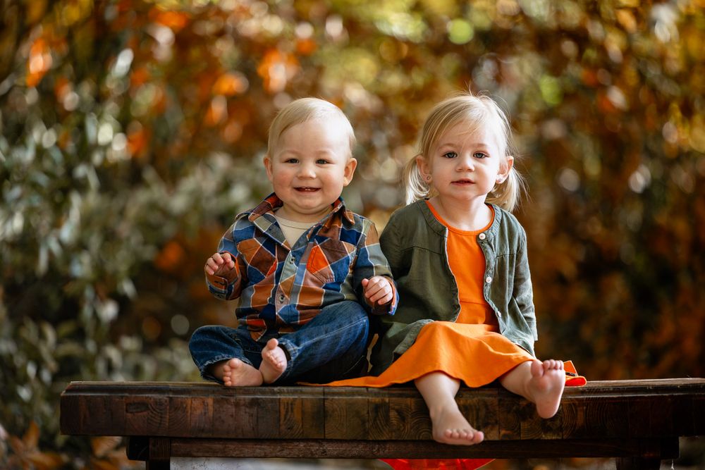 Two small children sit together on a wooden bench amid fall foliage.