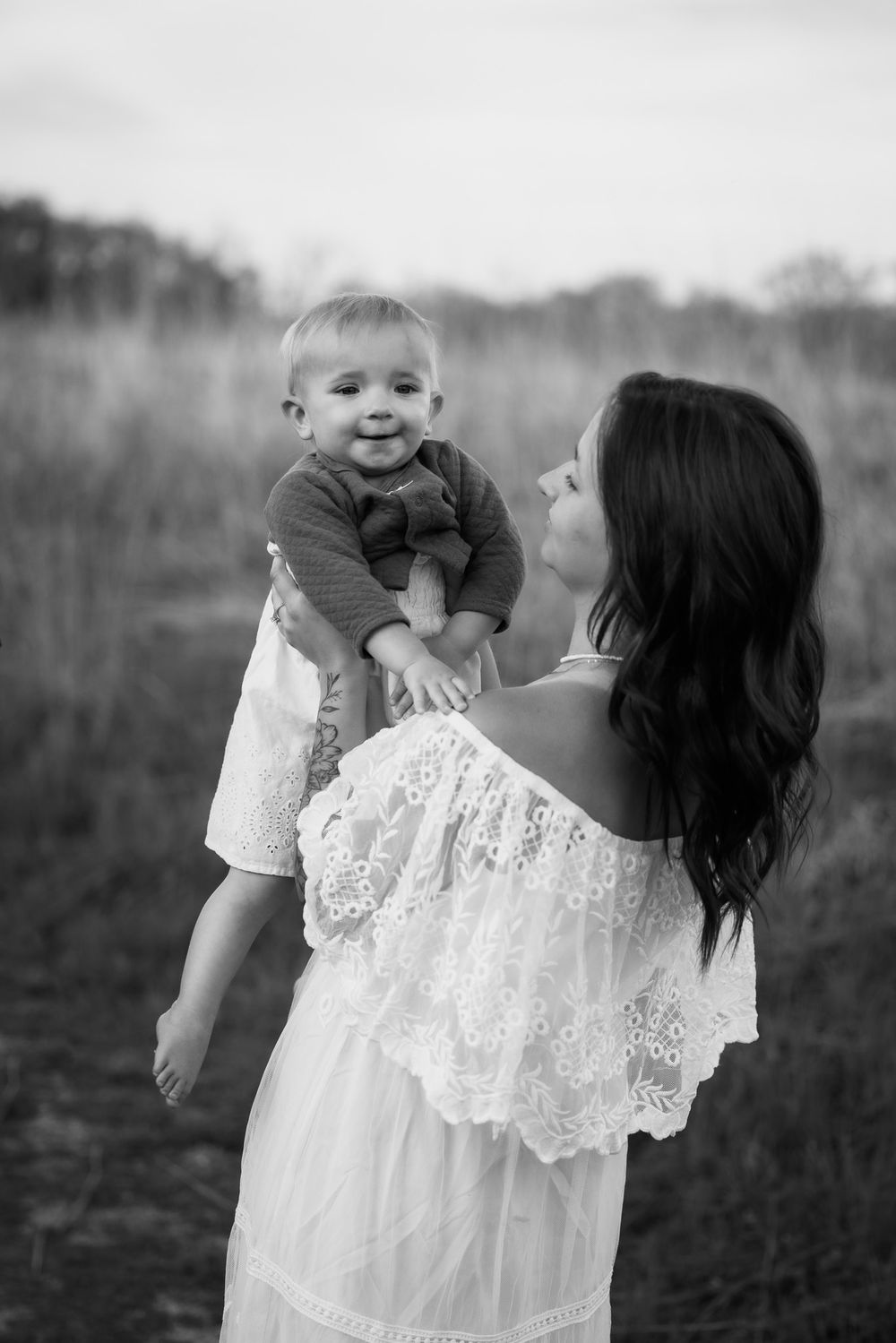Mother in white lace dress holding smiling baby outdoors in black and white portrait.