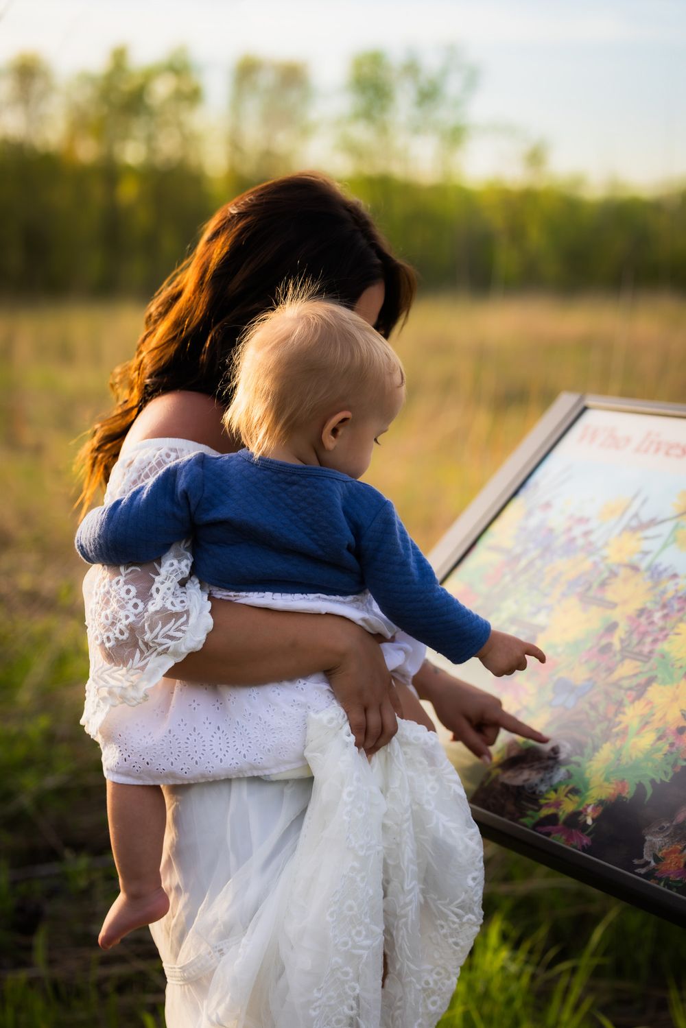Mother in white dress showing painting to small child in outdoor setting at sunset.