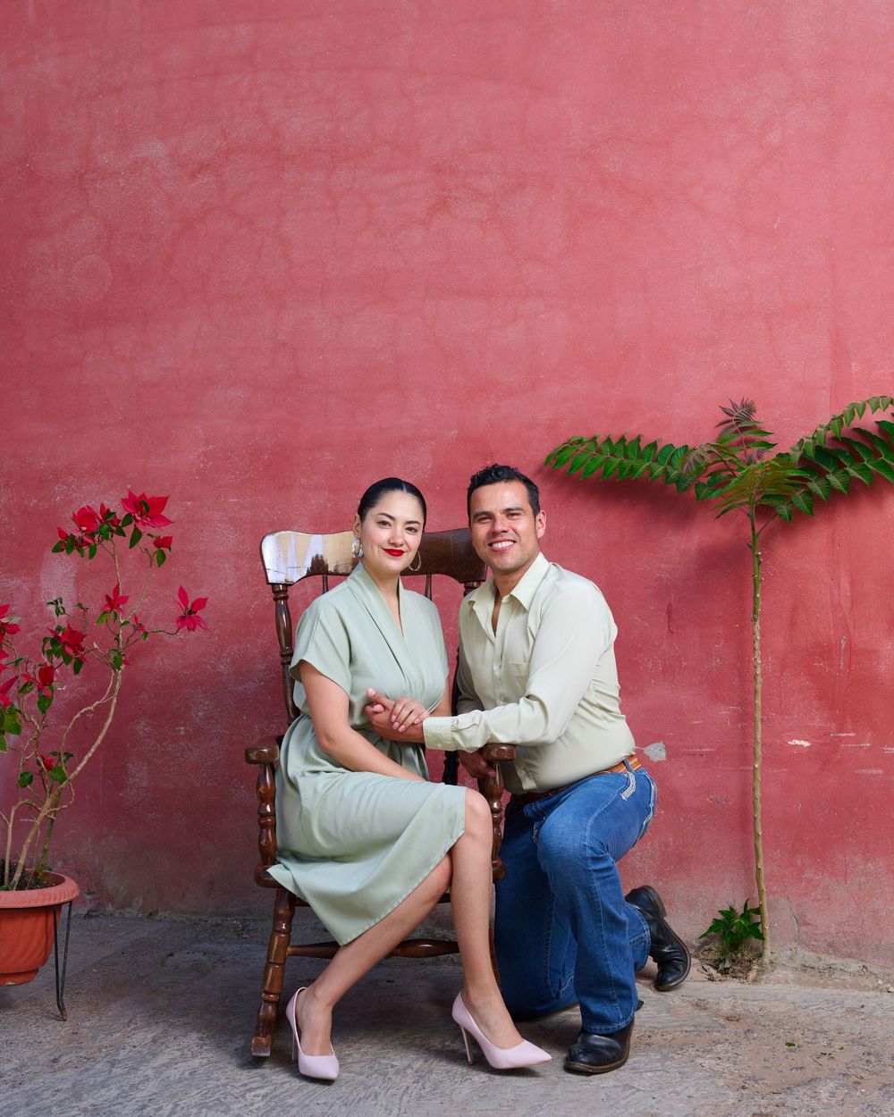 A couple poses against a coral-pink wall with flowering plants and greenery, creating a romantic garden atmosphere.