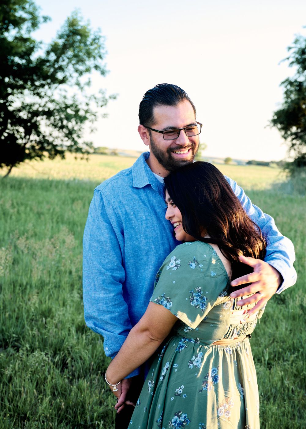 A tender embrace shared between a couple standing in a sunny meadow with green grass and trees in the background.