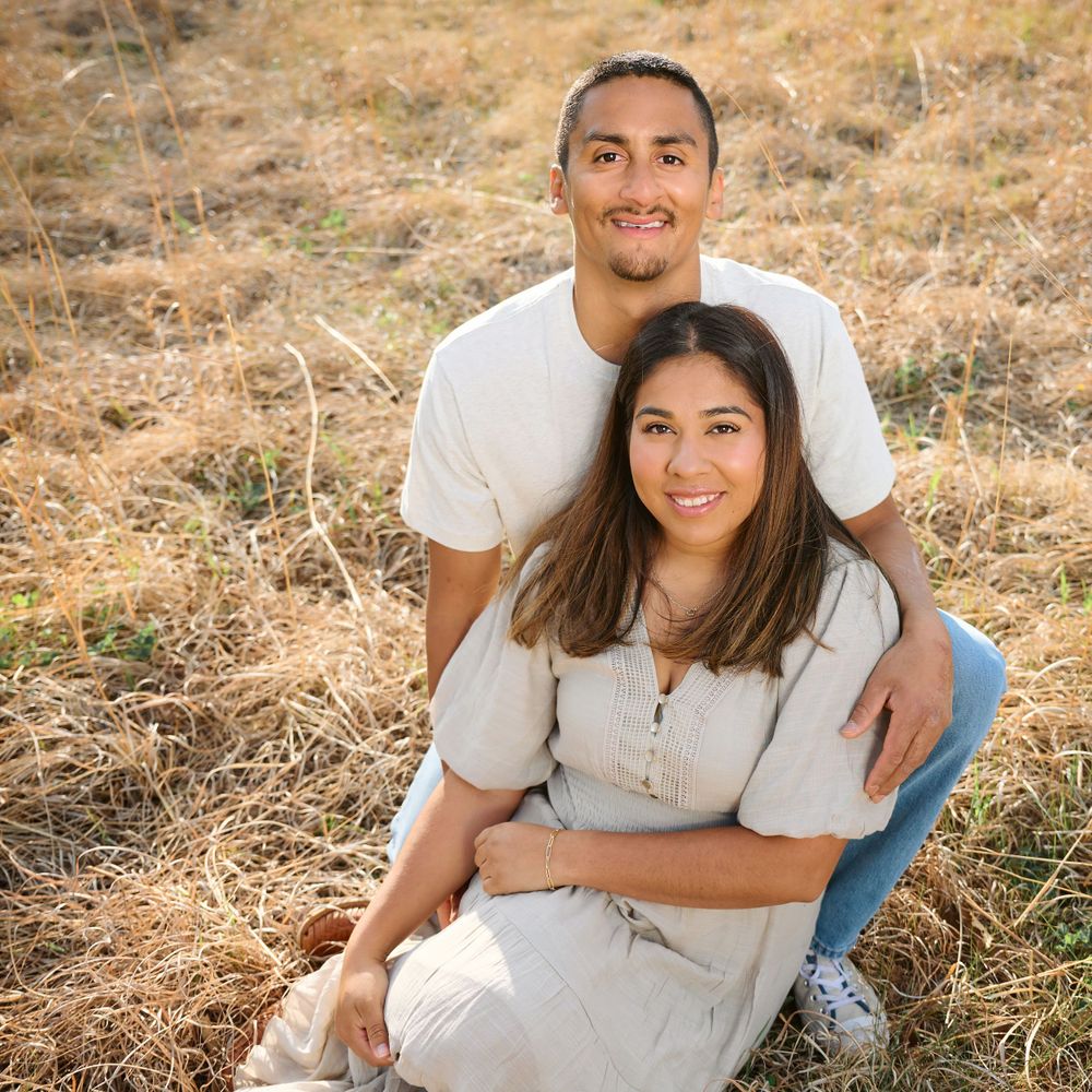 Couple in white outfits poses for engagement photo in field of dry grass.