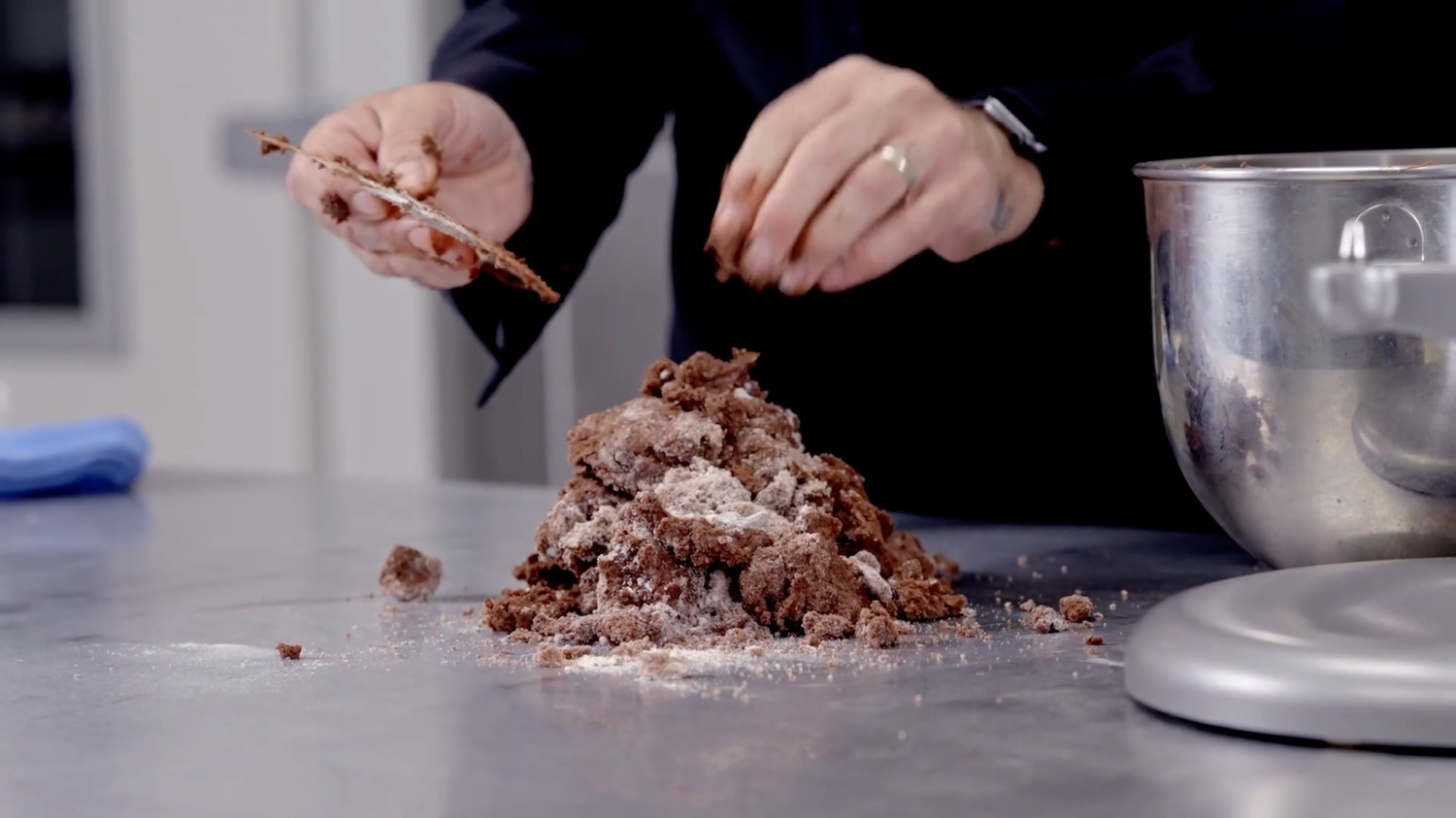 Hands breaking apart chocolate mixture on dark surface next to silver mixing bowl.