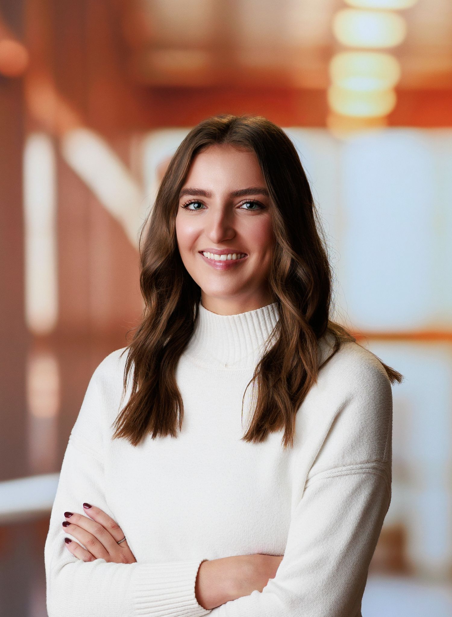Young woman in white turtleneck sweater smiling with arms crossed in a modern office setting for headshot photos