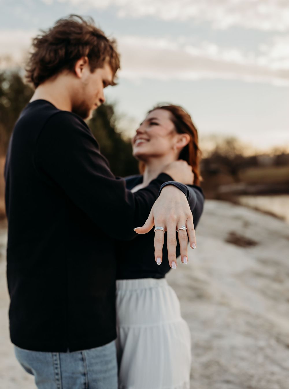 A couple embraces on a beach at sunset, showing off an engagement ring in a series of romantic photos.