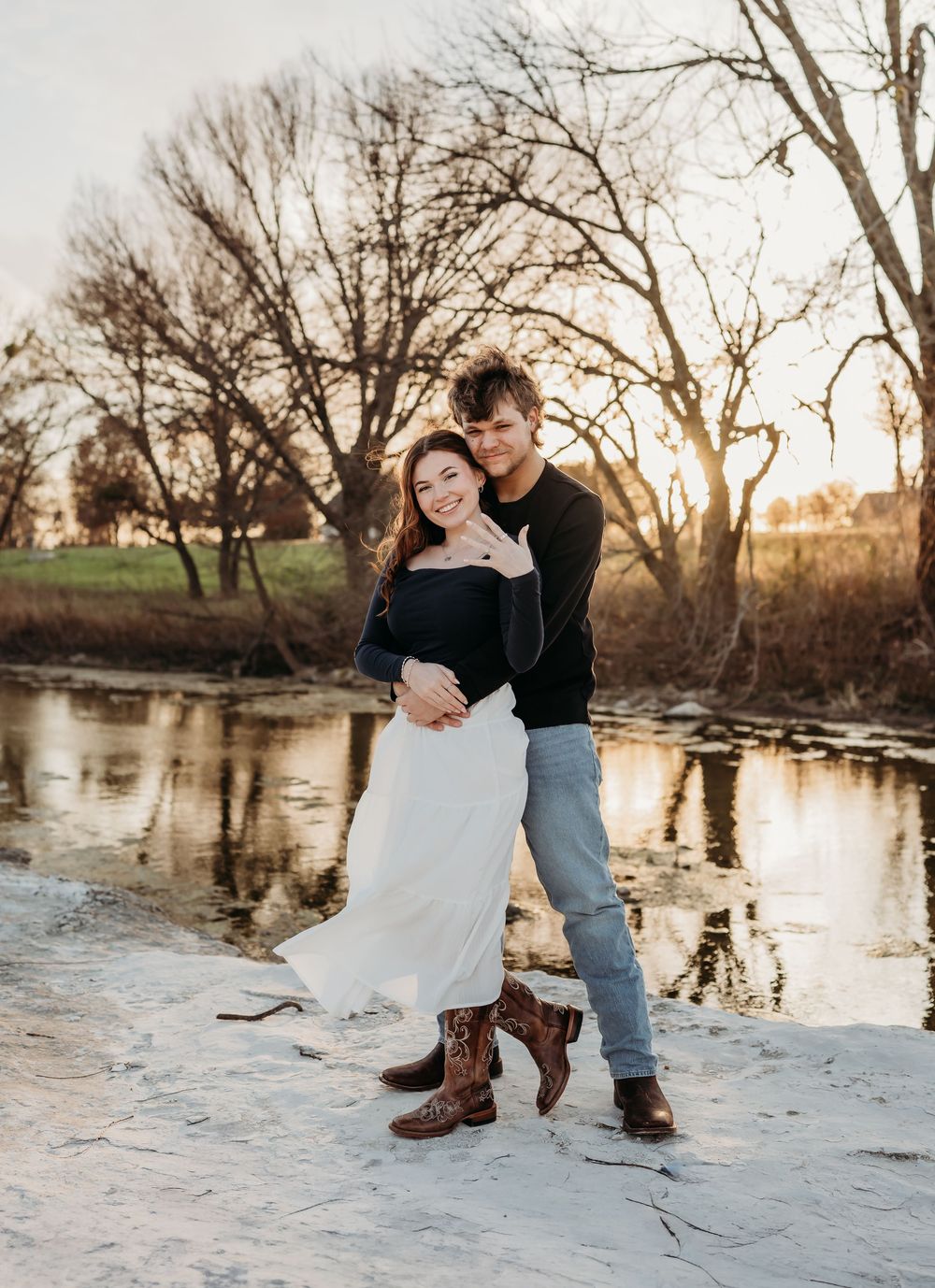 A couple shares a romantic moment on snowy ground by a river at sunset with bare winter trees in the background.