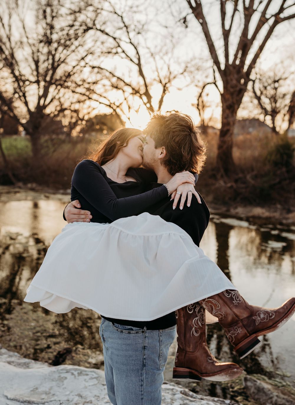 A romantic sunset moment by a pond with brown cowboy boots and a flowing white skirt against bare winter trees.