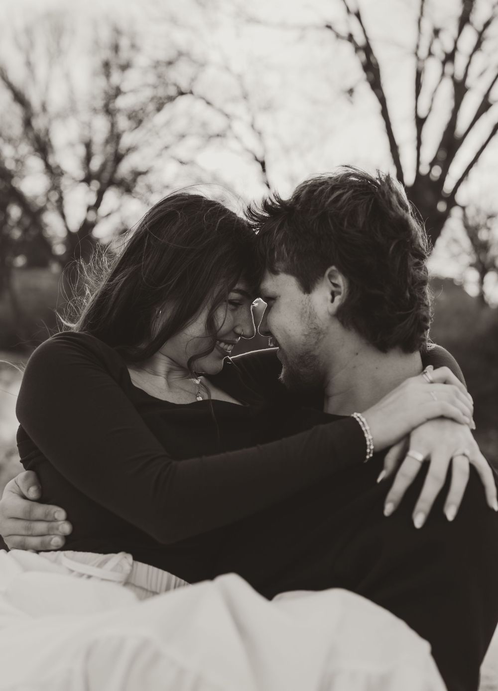 An intimate black and white portrait of a couple sharing a tender moment outdoors with bare trees in background.