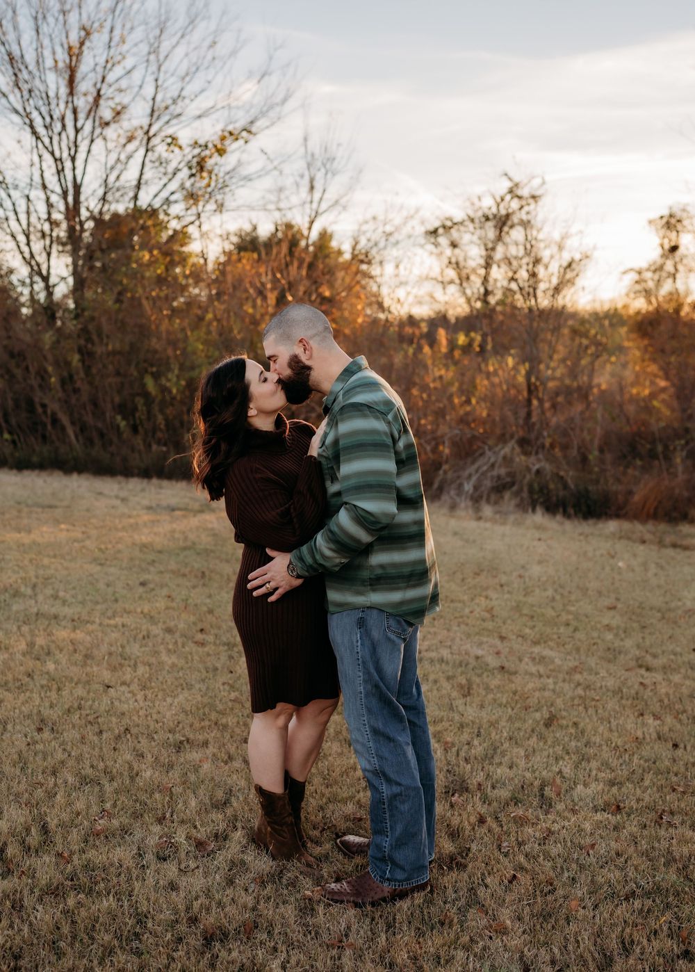 A romantic couple embraces in a field during golden hour with autumn trees in the background.
