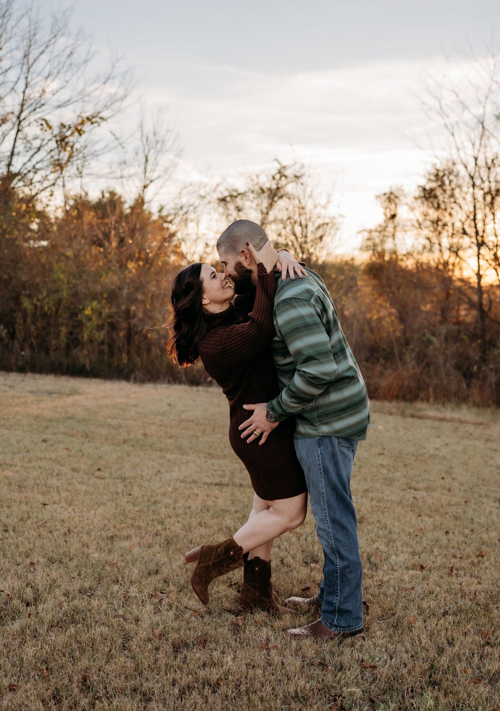 A couple shares an intimate moment in a rustic field at sunset with fall foliage behind them.