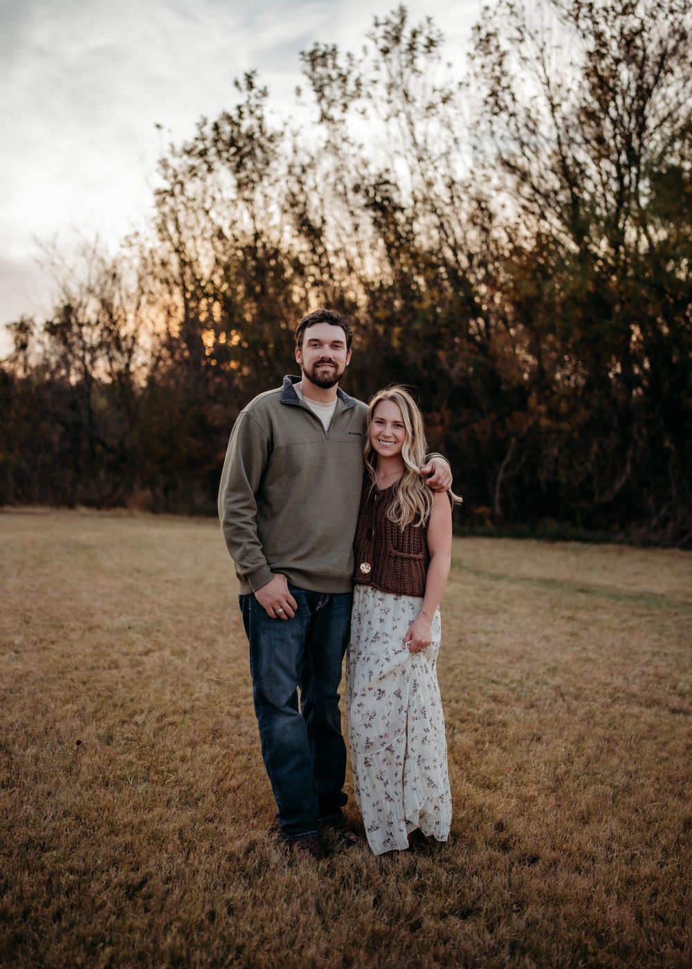 A couple stands together in a grassy field at sunset with trees silhouetted in the background.