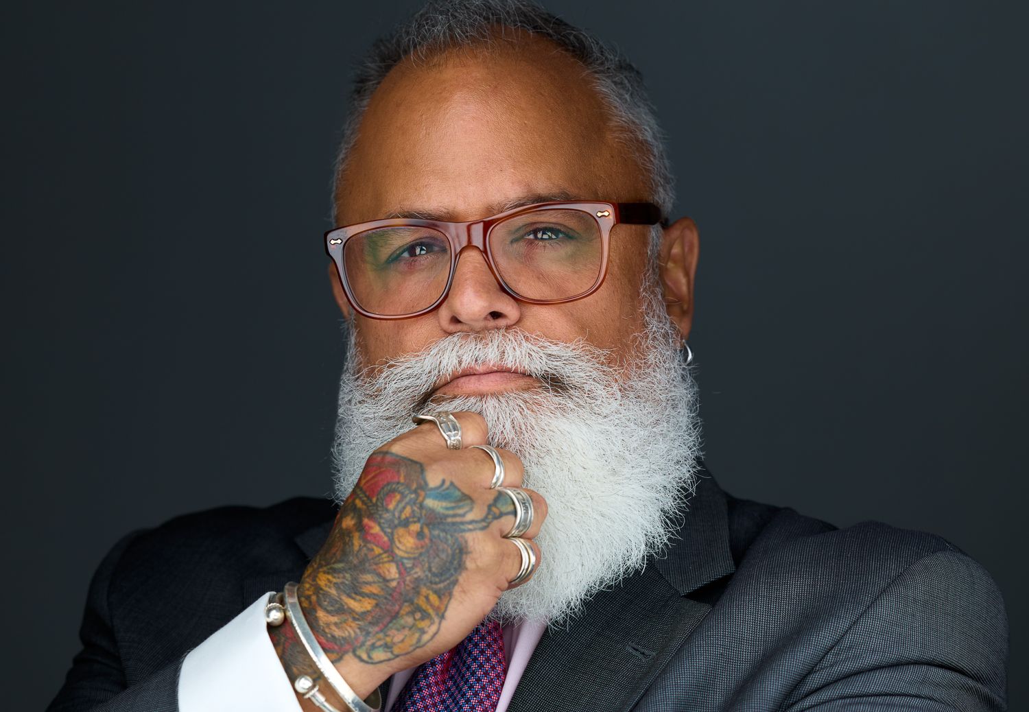 Professional in gray suit and glasses with white beard and tattooed hand photographed against dark background.