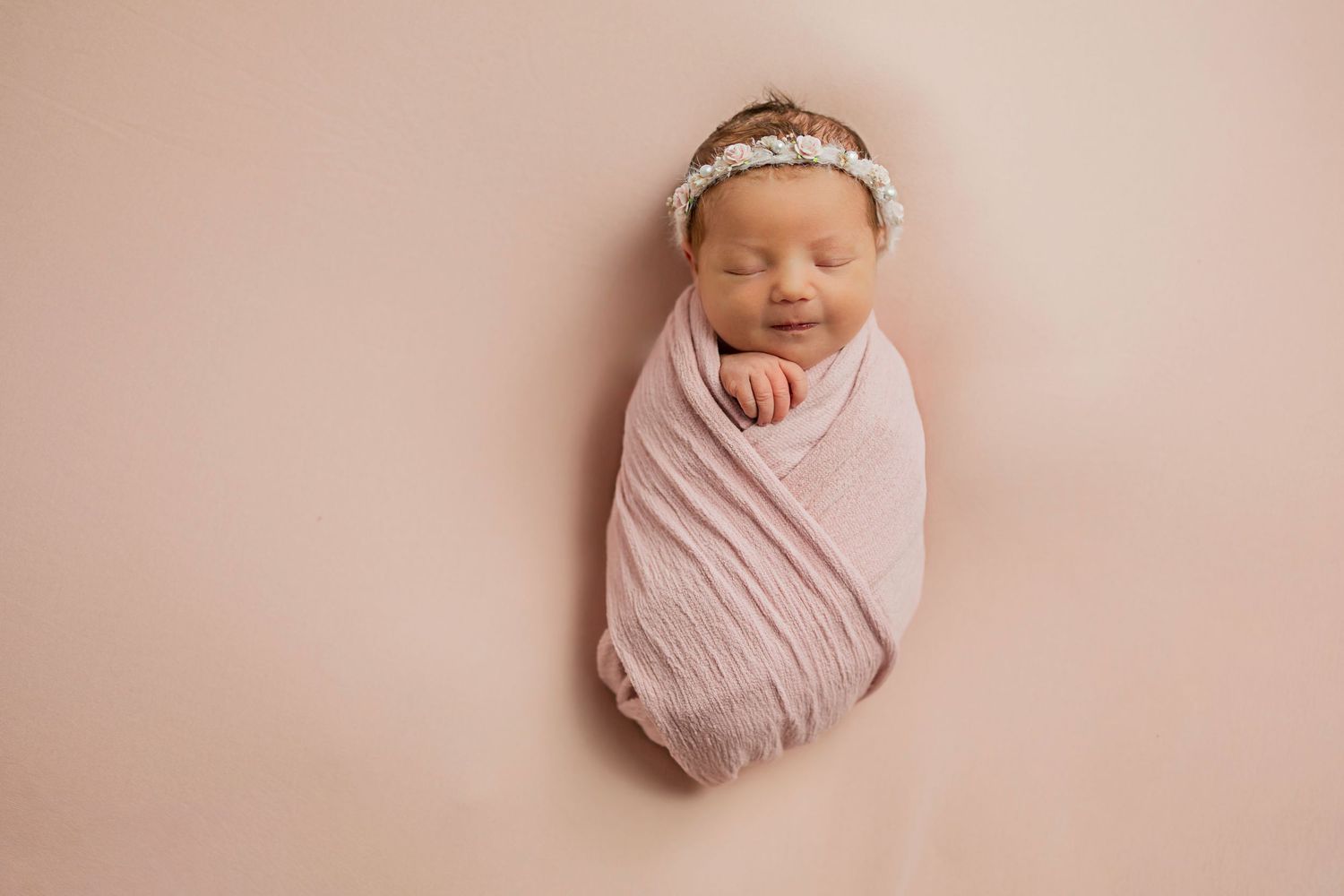Newborn baby girl swaddled in pink wrap with floral headband sleeps on pink background.