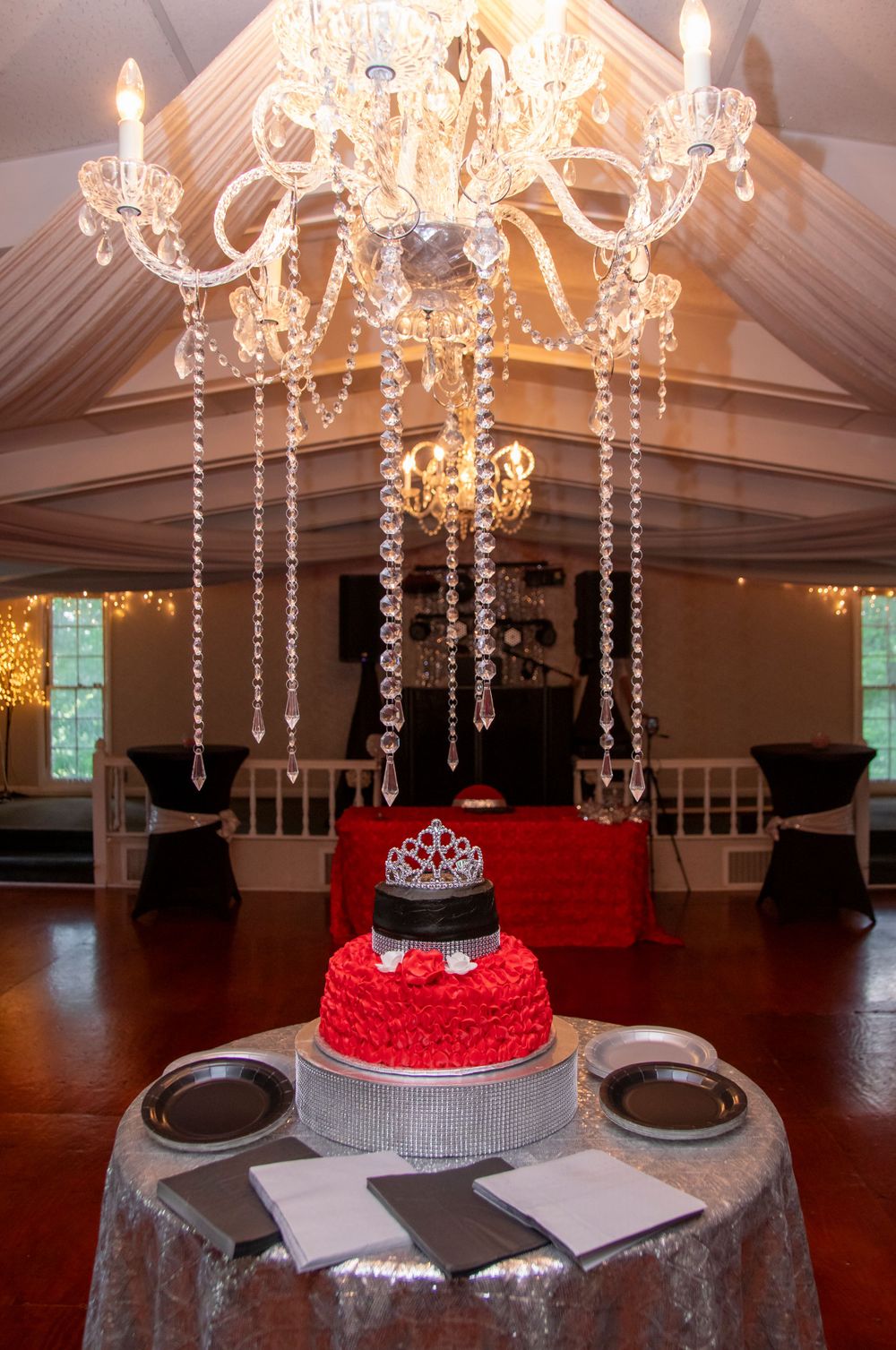 Elegant crystal chandelier hanging above red and silver tiered cake with crown topper.