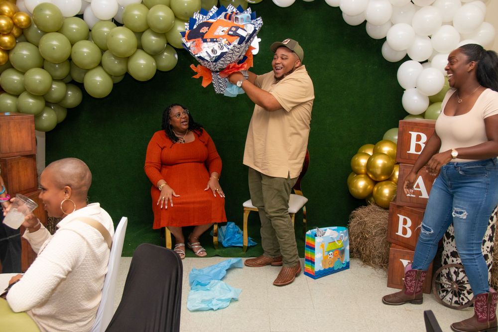 People gather at a baby shower celebration with green and white balloon decorations and a 'BABY' sign in the background.
