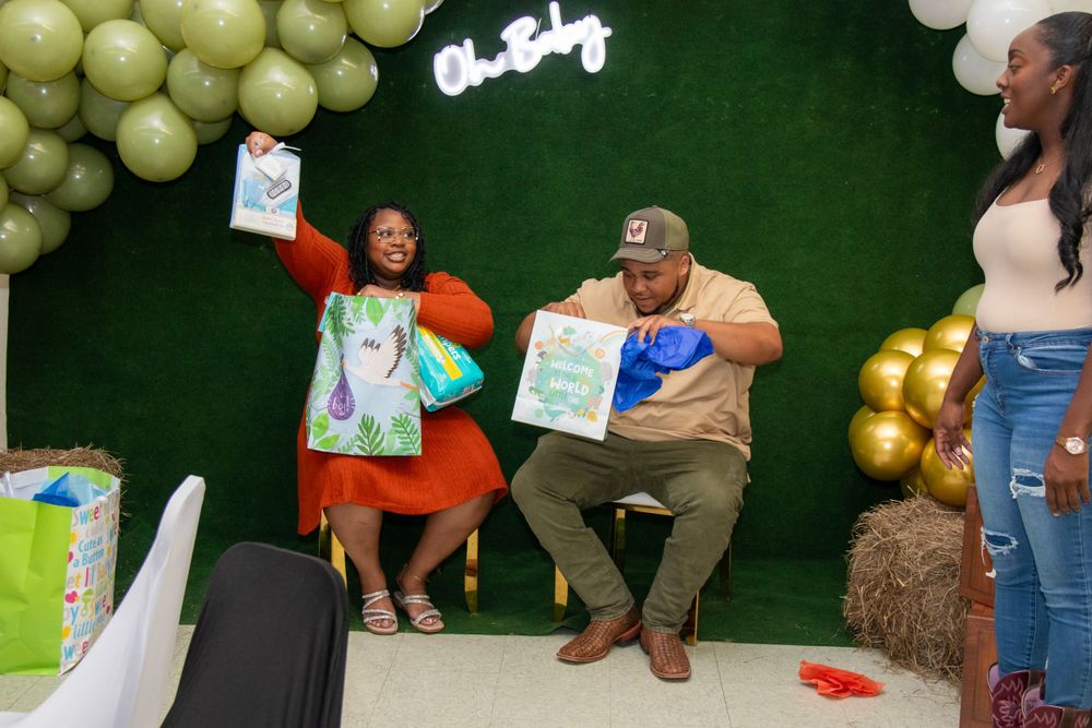 Baby shower celebration with guests holding gift boxes against green backdrop with balloon arch.