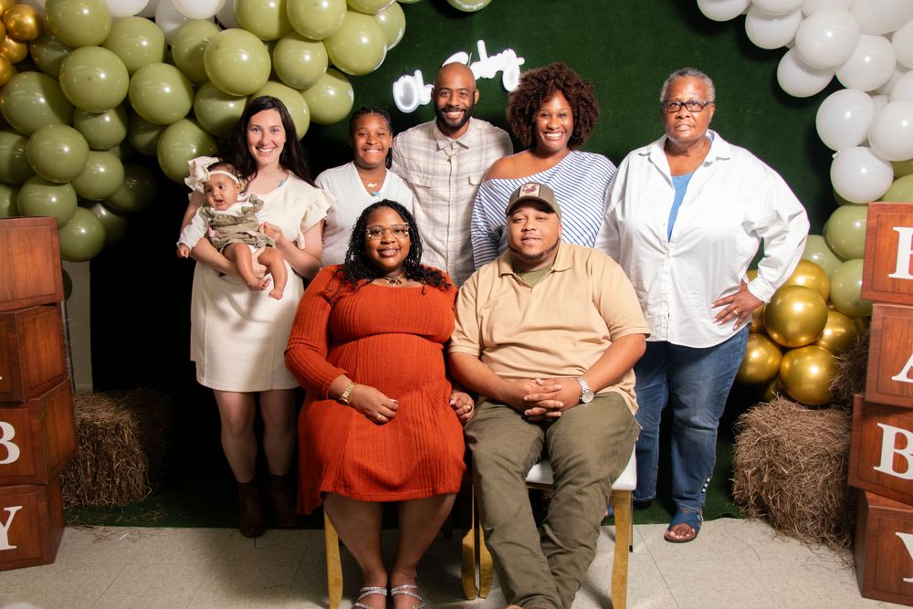 Group photo against green backdrop with gold and white balloon arch decorations and rustic wooden accents.