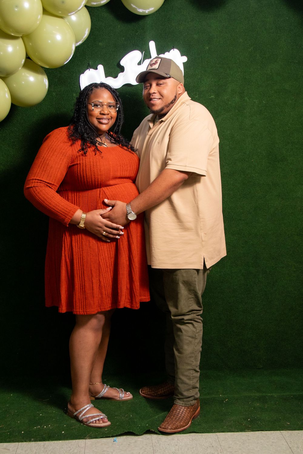 Expectant couple poses against green backdrop with yellow balloons during baby shower photoshoot.