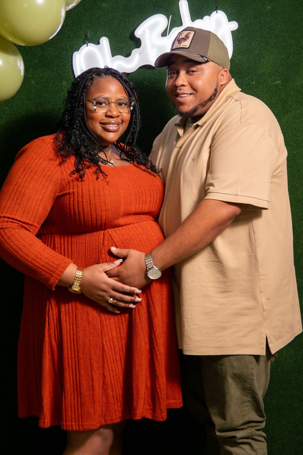 Couple poses together for a maternity photo against a green backdrop with gold balloons, the woman wearing a rust-colored dress.