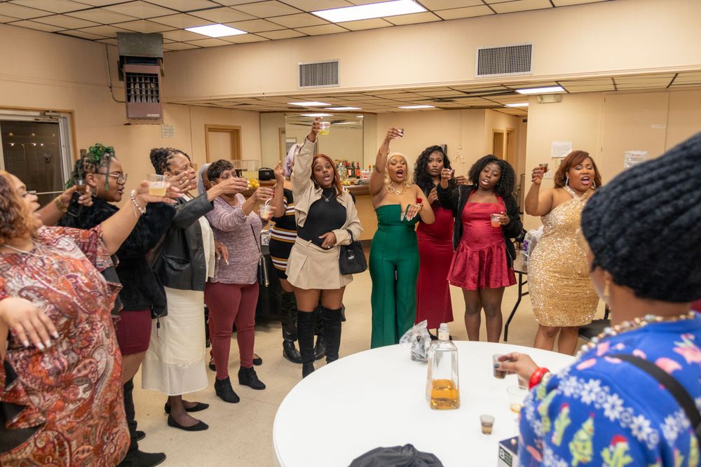 A group of people celebrate together at an indoor party, raising their hands in excitement by a table with drinks.