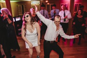 Guests dancing energetically at a wedding reception, woman in white dress with sunglasses and man in white shirt striking fun poses.