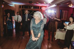 Elderly woman dancing energetically at a wedding reception on a wooden dance floor surrounded by guests.