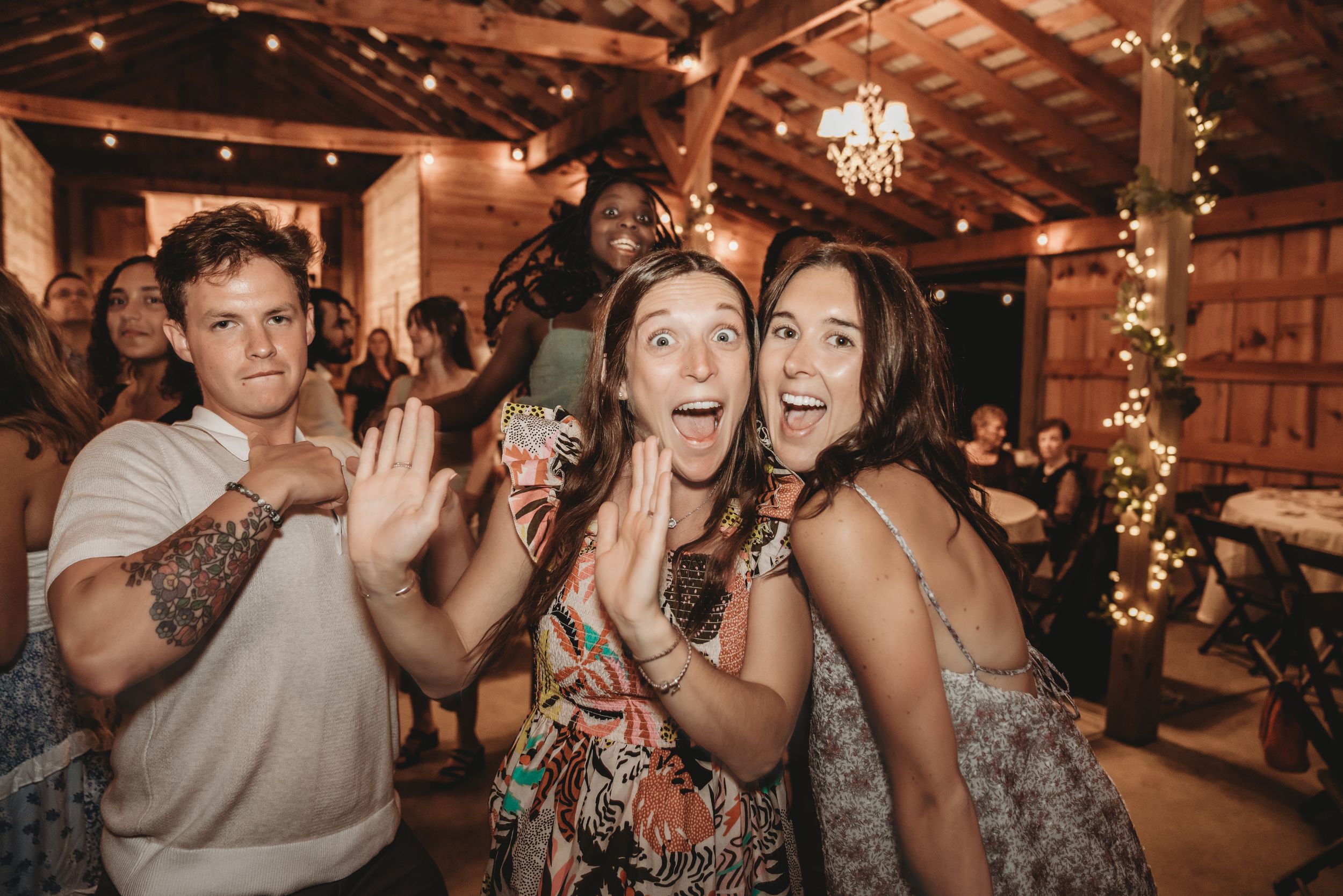 Excited guests react with surprise and joy at a rustic barn party with string lights in the background.