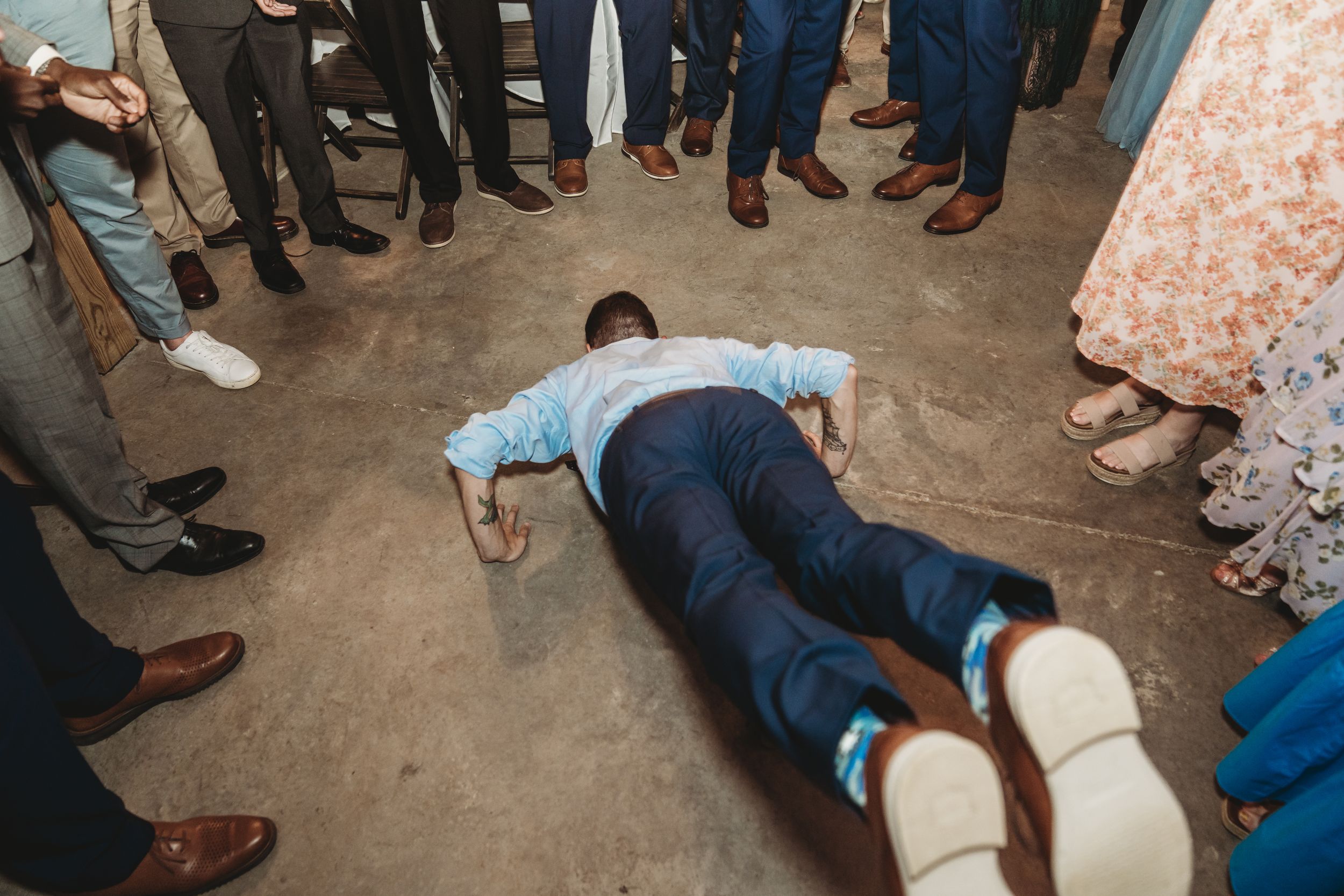 Man lying face down on concrete floor surrounded by wedding guests in formal attire at a reception.