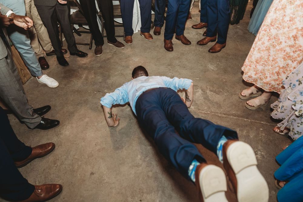 Man lying face down on concrete floor surrounded by wedding guests in formal attire at a reception.