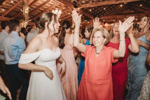 Bride in white off-shoulder gown dancing joyfully with guests at a rustic wedding reception.