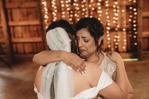 A bride in a white gown and veil shares an emotional hug with her mother at a rustic barn wedding reception.