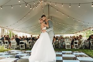 Couple sharing first dance under string lights in elegant outdoor wedding tent with guests seated around checkered dance floor.