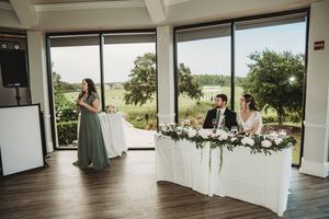 Bridesmaid giving a speech at an elegant wedding reception with floor-to-ceiling windows overlooking a green landscape.