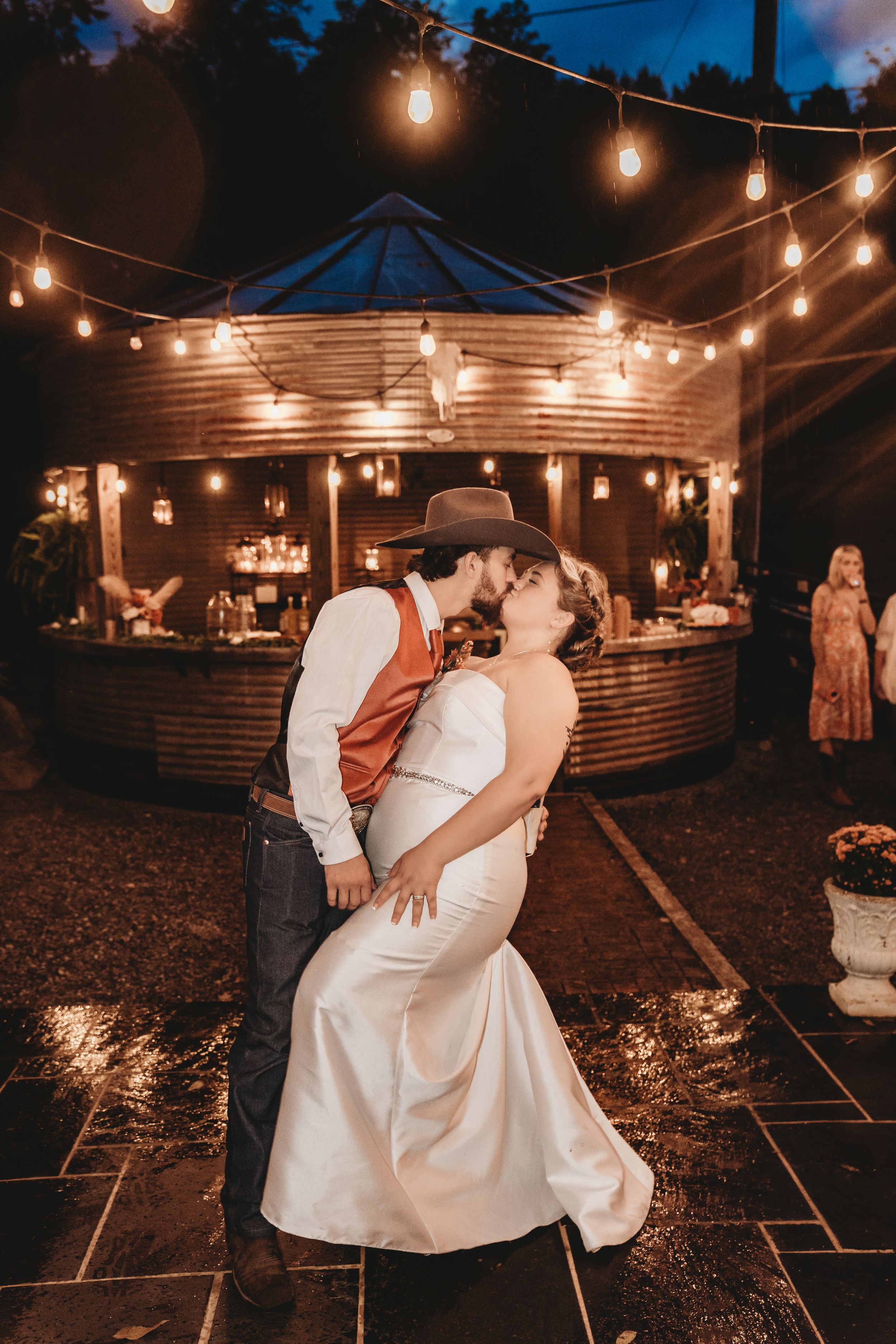 Bride and groom share a romantic kiss under string lights at an outdoor evening wedding reception.