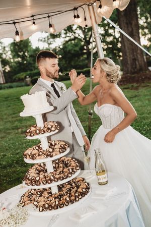Bride and groom feeding each other cake at outdoor wedding reception with tiered donut display and champagne bottle.