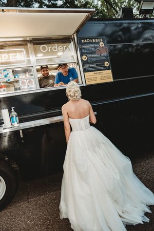 Bride in strapless ball gown ordering from Taco Bros food truck at outdoor wedding reception.