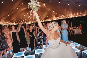 Bride in white gown tossing bouquet to guests on illuminated dance floor under tent with string lights.