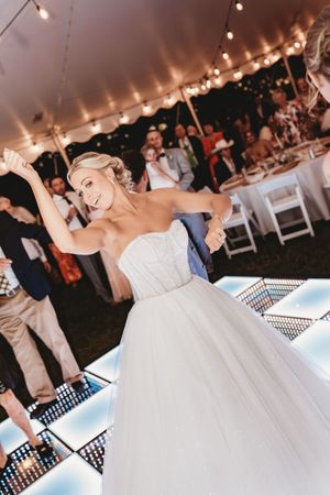 Smiling bride in strapless tulle gown dancing on illuminated floor under string lights at wedding.